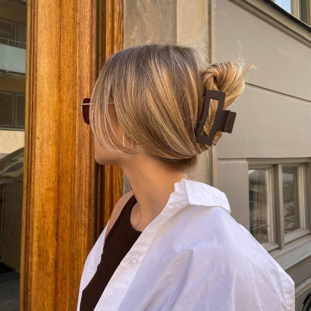 Brun hårklämma i knut – stylad i blont hår på modell i solglasögon och vit skjorta utanför entré
